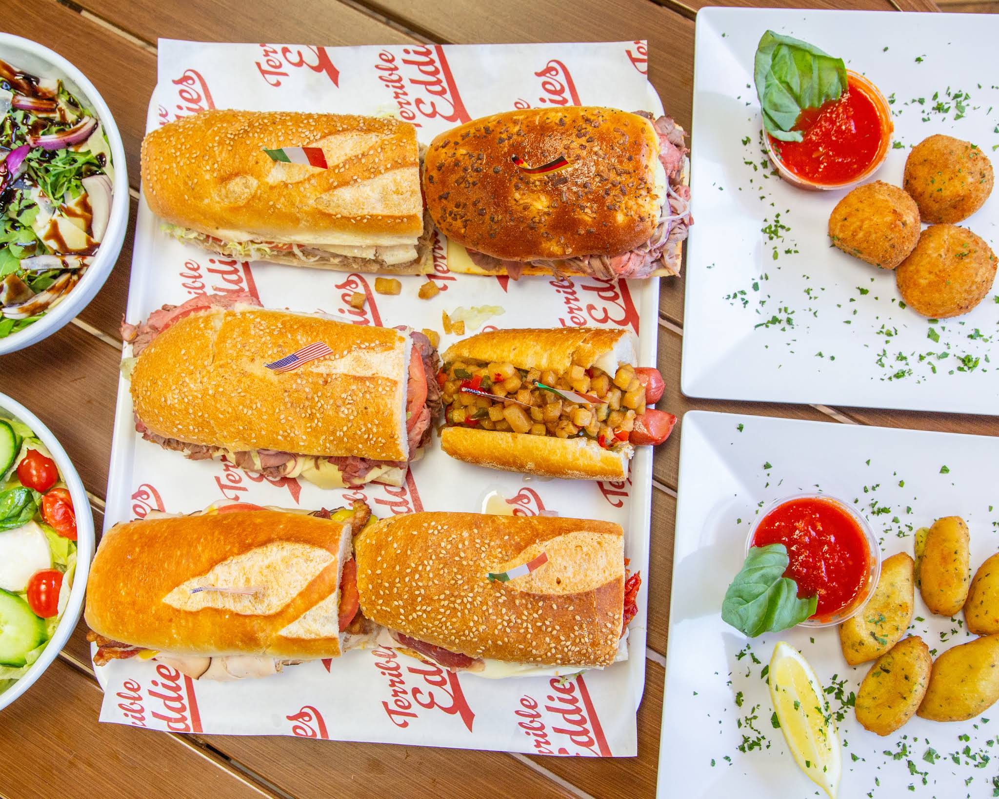 An overhead spread of Terrible Eddie’s sandwiches, salads and golden sides on branded paper